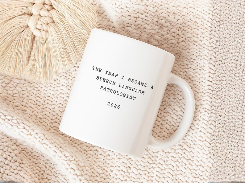 Op de afbeelding: Witte keramische mok met de tekst "THE YEAR I BECAME A SPEECH LANGUAGE PATHOLOGIST 2026". De mok staat op een cr&egrave;mekleurig gebreid oppervlak met een beige macram&eacute; decoratie.