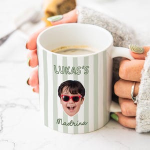 Puede incluir: Una taza de cer&aacute;mica blanca con rayas verticales de color verde salvia, un retrato fotogr&aacute;fico de un ni&ntilde;o con gafas de sol rojas y el texto impreso LUKAS'S y Madrina en fuente verde oscuro. La taza tiene un asa y contiene un l&iacute;quido oscuro.