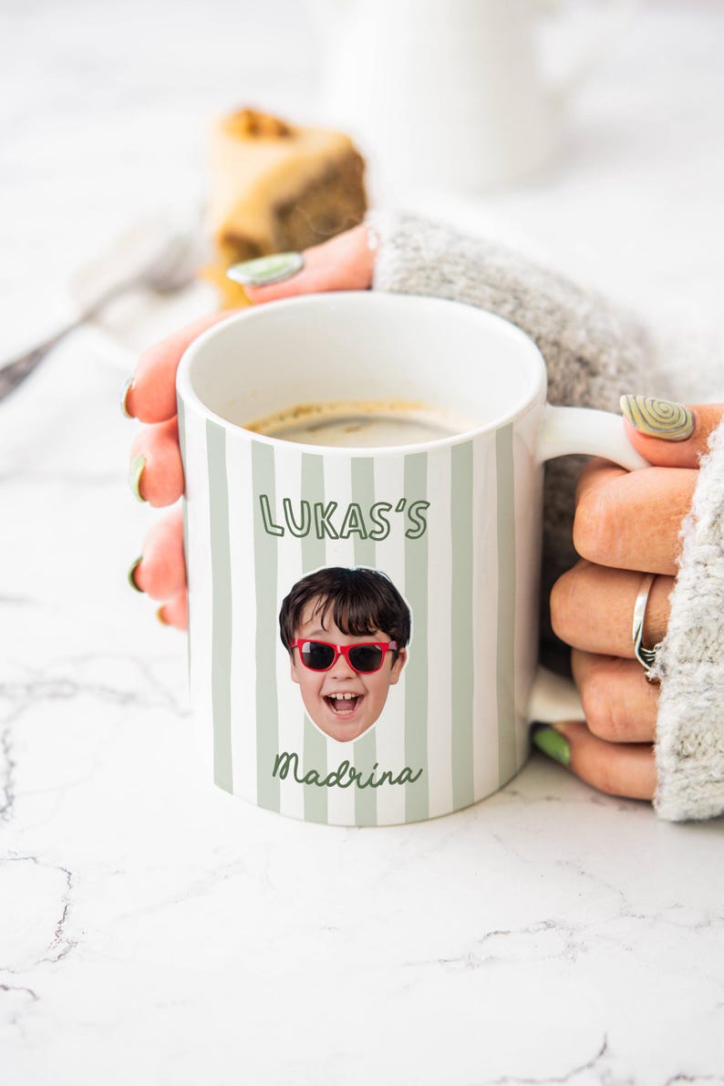 Puede incluir: Una taza de cer&aacute;mica blanca con rayas verticales de color verde salvia, un retrato fotogr&aacute;fico de un ni&ntilde;o con gafas de sol rojas y el texto impreso LUKAS'S y Madrina en fuente verde oscuro. La taza tiene un asa y contiene un l&iacute;quido oscuro.
