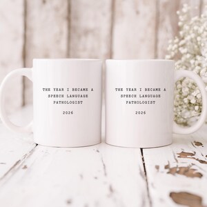 Op de afbeelding: Twee witte keramische mokken met zwarte tekst die luidt: "THE YEAR I BECAME A SPEECH LANGUAGE PATHOLOGIST 2026". De mokken staan op een wit houten oppervlak met een vage achtergrond en een takje witte bloemen.