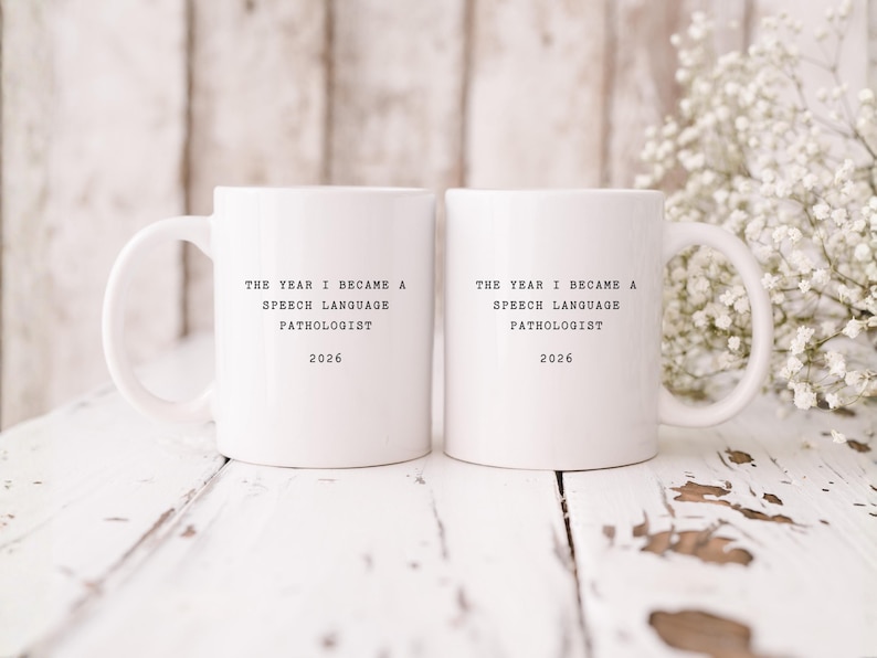 Op de afbeelding: Twee witte keramische mokken met zwarte tekst die luidt: "THE YEAR I BECAME A SPEECH LANGUAGE PATHOLOGIST 2026". De mokken staan op een wit houten oppervlak met een vage achtergrond en een takje witte bloemen.