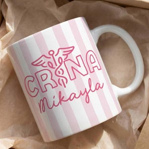 Pu&ograve; includere: Una tazza in ceramica bianca con strisce verticali rosa e un manico bianco. La tazza presenta la scritta "CRNA Mikayla" in rosa, insieme a un simbolo medico. La tazza &egrave; presentata all'interno di una scatola di cartone marrone.