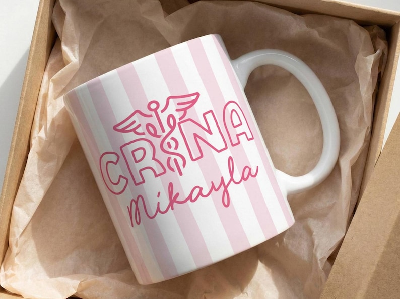 Pu&ograve; includere: Una tazza in ceramica bianca con strisce verticali rosa e un manico bianco. La tazza presenta la scritta "CRNA Mikayla" in rosa, insieme a un simbolo medico. La tazza &egrave; presentata all'interno di una scatola di cartone marrone.
