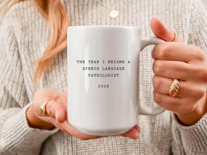 May include: White ceramic mug with the text "THE YEAR I BECAME A SPEECH LANGUAGE PATHOLOGIST 2026". The mug is held by a person wearing a gray sweater and gold rings.