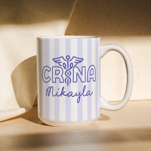 Pu&ograve; includere: Tazza in ceramica bianca con un motivo a righe blu e bianche. La tazza presenta il testo "CRNA Mikayla" in blu, insieme a un simbolo di caduceo. Ha un manico grande ed &egrave; adatta per bevande calde o fredde.