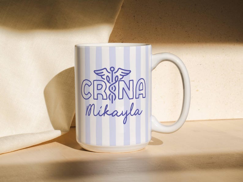 Pu&ograve; includere: Tazza in ceramica bianca con un motivo a righe blu e bianche. La tazza presenta il testo "CRNA Mikayla" in blu, insieme a un simbolo di caduceo. Ha un manico grande ed &egrave; adatta per bevande calde o fredde.