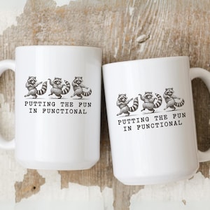 May include: Two white ceramic mugs with a black and white illustration of raccoons and the text "PUTTING THE FUN IN FUNCTIONAL". The mugs have large handles and are set against a rustic wooden background.