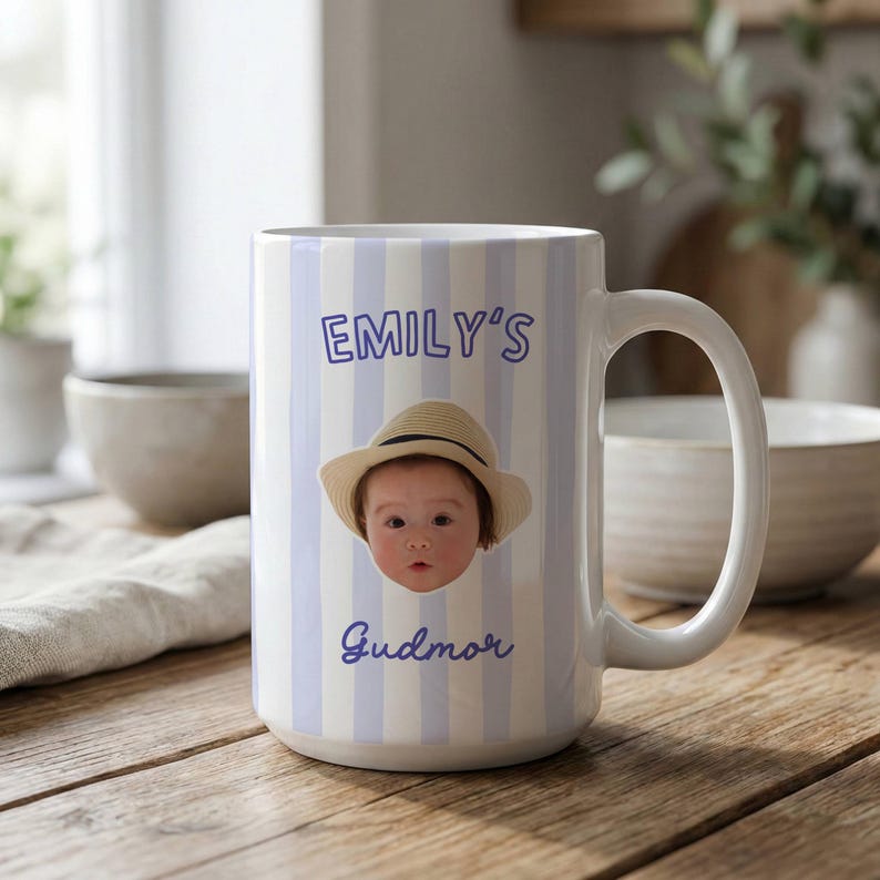Puede incluir: Una taza de cer&aacute;mica blanca con rayas verticales de color azul claro, una fotograf&iacute;a de un beb&eacute; con sombrero de paja y el texto morado "EMILY'S" sobre la imagen y "Gudmor" debajo, con un asa curva en el lado derecho.