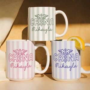 Pu&ograve; includere: Tre tazze in ceramica bianca con motivi a righe verticali verdi, rosa e blu. Ogni tazza presenta il testo "CRNA" con un simbolo medico e il nome "Mikayla". Una fetta di limone poggia su una tazza.