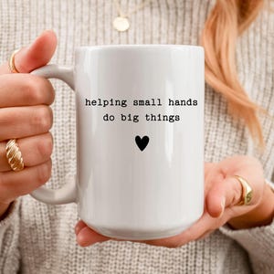 May include: White ceramic mug with the text "helping small hands do big things" and a black heart. The mug is held by a person wearing gold rings and a sweater.