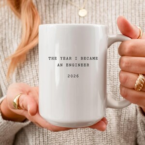 May include: A white ceramic mug with the text "THE YEAR I BECAME AN ENGINEER 2026" in black. The mug has a large handle and is held by a person. The mug is a gift for an engineer.