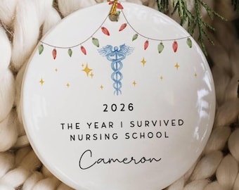 Op de afbeelding: Een rond wit keramieken ornament met een blauw caduceussymbool, gele sterren en een snoer met rode en groene kerstverlichting. De tekst luidt 2026, THE YEAR I SURVIVED NURSING SCHOOL, en de naam Cameron in een zwart cursief lettertype.