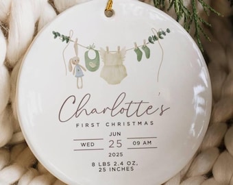 Op de afbeelding: Een rond wit keramieken ornament met een gouden ophangkoord. Het ontwerp toont een waslijn met een knuffel, slabbetje, rompertje en sokjes. Tekst: Charlotte's FIRST CHRISTMAS JUN WED 25 09 AM 2025 3,7 kg, 63,5 cm.
