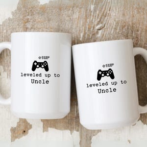 May include: Two white ceramic mugs with black handles. The mugs have a black video game controller graphic and the text "Level Up to Uncle".