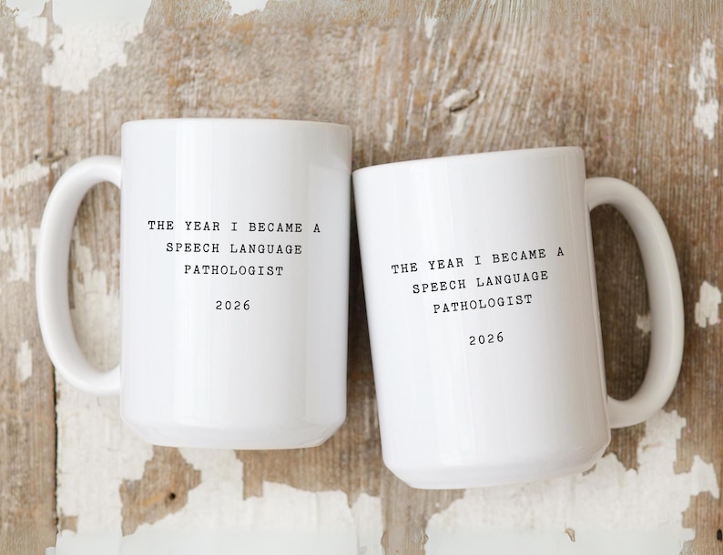 Op de afbeelding: Twee witte keramische mokken met zwarte tekst. De tekst op de mokken luidt "THE YEAR I BECAME A SPEECH LANGUAGE PATHOLOGIST 2026". De mokken hebben grote handvatten en staan tegen een houten achtergrond.