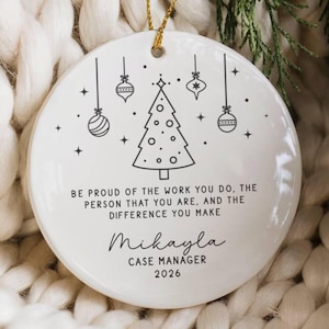 May include: A round white ceramic ornament featuring a black line art design of a Christmas tree and hanging baubles. The text reads: BE PROUD OF THE WORK YOU DO, THE PERSON THAT YOU ARE, AND THE DIFFERENCE YOU MAKE, Mikayla, CASE MANAGER, 2026.