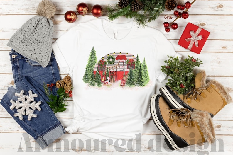 K&ouml;nnte beinhalten: Wei&szlig;es T-Shirt mit einer Aquarellillustration eines roten Wohnwagens, der f&uuml;r Weihnachten geschm&uuml;ckt ist und von gr&uuml;nen Tannenb&auml;umen umgeben ist. Das Bild befindet sich auf einem rustikalen Holzgrund, mit Winteraccessoires wie einer grauen M&uuml;tze, Jeans und Stiefeln.