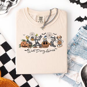 May include: Cream-colored t-shirt featuring a Halloween-themed graphic of Disney characters dressed as skeletons, with pumpkins and the text "Walt Disney World." The shirt is paired with denim shorts, a silver chain, and Halloween decorations.