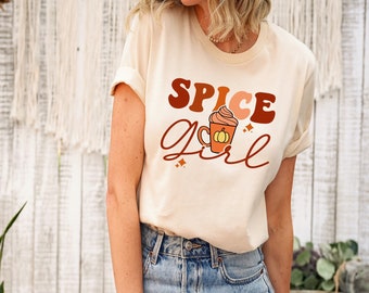 May include: Beige t-shirt with the words "SPICE Girl" in brown and orange lettering, featuring a graphic of a pumpkin spice latte. The shirt is a casual style, suitable for fall. The model is wearing blue jeans.