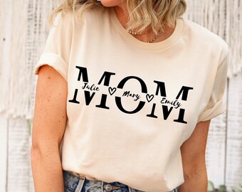 May include: A cream colored t-shirt with the word "MOM" printed in black. The word is divided into three sections with the names "Julie", "Mary", and "Emily" printed in black with a heart symbol between each name.