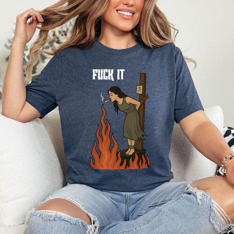May include: Navy blue t-shirt featuring the white text "FUCK IT". The graphic shows a woman tied to a stake, smoking a cigarette, with flames around her. The woman is wearing a dark green dress.