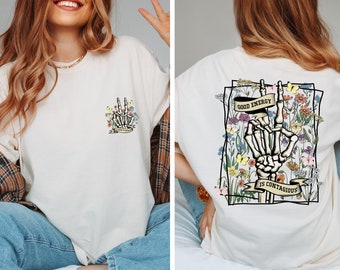 May include: Cream-colored t-shirt featuring a skeleton hand making a peace sign, surrounded by flowers and butterflies. The text "Good Energy Is Contagious" is displayed on the back. A smaller version of the design is on the front.