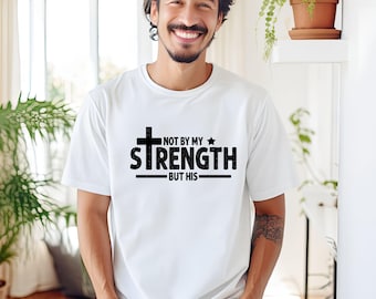 May include: White t-shirt with a black graphic design that says "Not by my strength but his" with a cross and a star.
