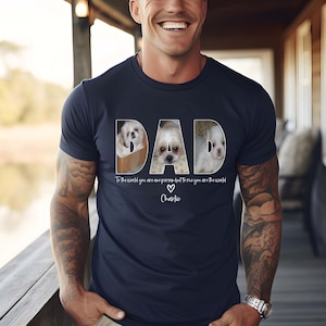 Pu&ograve; includere: Una maglietta blu navy con una grafica bianca della parola "DAD" composta da tre foto di cani. Il testo "To the world you are one person but to me you are the world" &egrave; sotto la grafica, con un cuore e il nome "Charlie" sotto il testo.