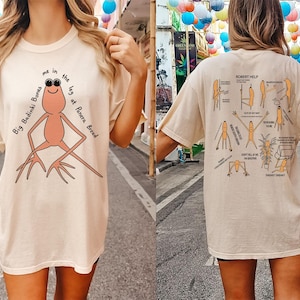 May include: Beige t-shirt with a cartoon frog wearing sunglasses and the text "Big Badinki Bones me in the leg at Panera Bread." The back of the shirt features a diagram with the text "ROBERT HELP."