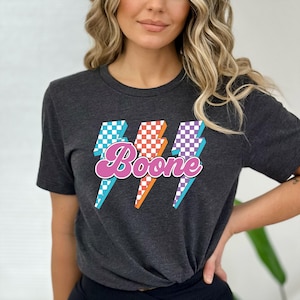 May include: Dark gray t-shirt featuring the name "Boone" in a pink, retro font, with three lightning bolt graphics in blue, orange, and purple checkered patterns. The shirt has short sleeves and a crew neck.