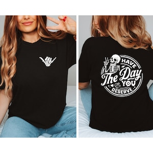May include: Black t-shirt with a white skeleton hand making a shaka sign on the front and a white circle on the back with a skeleton holding a peace sign and the text "Have The Day You Deserve."
