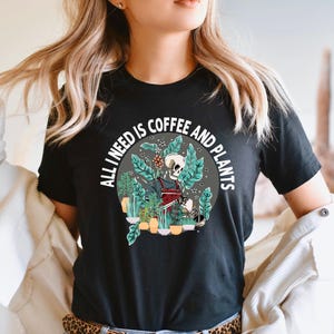 May include: Black t-shirt with a graphic of a skeleton holding a coffee cup and watering plants. The text "All I need is coffee and plants" is written in a circle around the skeleton.