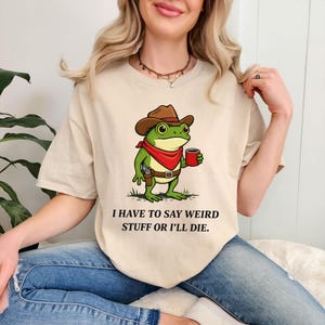 May include: Beige t-shirt featuring a cartoon frog wearing a cowboy hat, red bandana, and holding a mug. The frog has a gun holster. Text on the shirt reads: "I HAVE TO SAY WEIRD STUFF OR I'LL DIE."