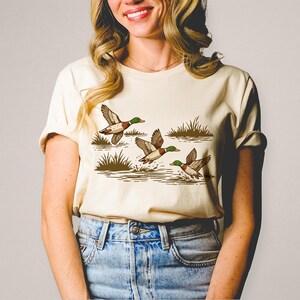 May include: Cream-colored t-shirt featuring a graphic of three ducks in flight over water and grass. The ducks have brown and white bodies with green heads. The design is in a vintage, outdoorsy style.