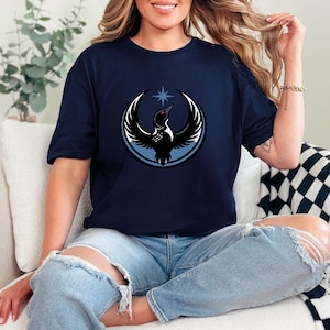 May include: Navy blue t-shirt featuring a circular graphic of a black bird with white wings, a red eye, and a blue star. The bird is set against a blue and white circular background. The shirt is made of a soft material.