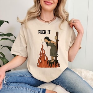 May include: Beige t-shirt featuring the text "FUCK IT" in black. The graphic shows a woman tied to a stake, smoking a cigarette, with flames around her. The woman is wearing a dark dress.