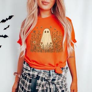 May include: Orange t-shirt featuring a beige ghost design in a field of flowers with bees. The ghost has two black oval eyes. The shirt is tied at the waist and has short sleeves.