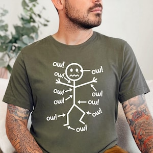 May include: Olive green t-shirt featuring a white stick figure with the word "ow!" pointing to various body parts. The stick figure has a distressed expression, with multiple instances of "ow!" around it.