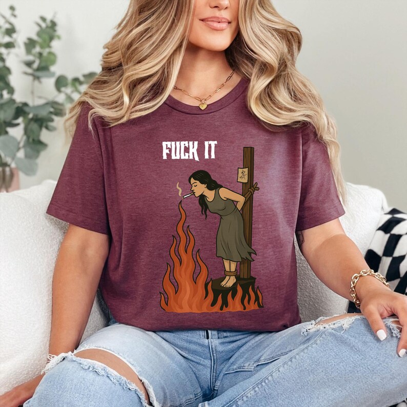 May include: Burgundy short-sleeved t-shirt featuring the text "FUCK IT" above a graphic of a person tied to a stake, smoking, with flames below. The graphic is in shades of brown, orange, and black.