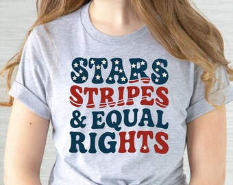 May include: A light gray t-shirt with the words "STARS STRIPES & EQUAL RIGHTS" in navy blue, red, and white. The word "STARS" has white stars on a navy blue background. The shirt is a crew neck with rolled-up sleeves.