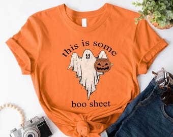May include: Orange short-sleeved t-shirt with a ghost holding a jack-o-lantern bucket. The text "this is some boo sheet" is printed on the shirt.
