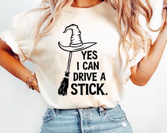 May include: Cream-colored t-shirt with a black graphic of a witch's hat and broom. The text "YES I CAN DRIVE A STICK" is printed below the hat and broom. The shirt is made of a soft material.