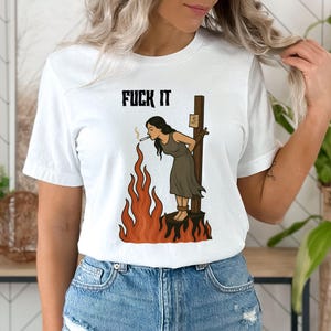May include: White t-shirt featuring the text "FUCK IT" above a graphic of a person tied to a stake, smoking, with flames below. The person is wearing a brown dress. The graphic is in black, brown, and orange.