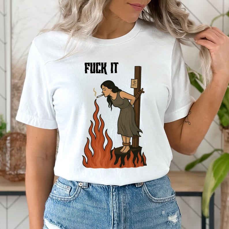 May include: White t-shirt featuring the text "FUCK IT" above a graphic of a person tied to a stake, smoking, with flames below. The person is wearing a brown dress. The graphic is in black, brown, and orange.