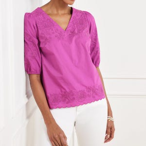 May include: A magenta blouse with a V-neck and short sleeves. The blouse has embroidered floral patterns on the neckline, sleeves, and hem. The hem has a scalloped edge. The blouse is a vibrant shade of pink.