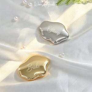 Peut inclure: Deux miroirs de poche aux formes irrégulières et liquides avec une finition polie. Un miroir est argenté et gravé du prénom Stacy, tandis que l'autre est doré et gravé du mot Bride dans une police d'écriture cursive.