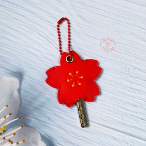 May include: Red leather keychain shaped like a cherry blossom with a gold key attached. The keychain has yellow stitching and a gold ring.