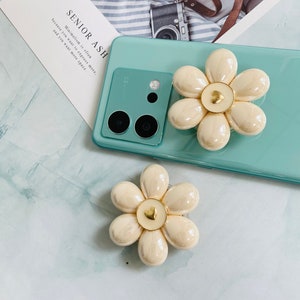 May include: A light blue phone case with a white flower-shaped phone grip. The grip has a gold heart in the center. The phone case has a camera lens on the back.