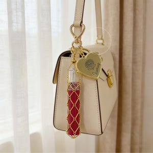 Könnte beinhalten: Ein goldfarbener Lippenstifthalter mit rotem Lippenstift, befestigt an einer cremefarbenen Handtasche. Der Lippenstifthalter ist in einem goldenen Netzdesign eingefasst. Ein goldfarbener herzförmiger Anhänger mit floralem Design und dem Wort "Icarus" ist ebenfalls angebracht.