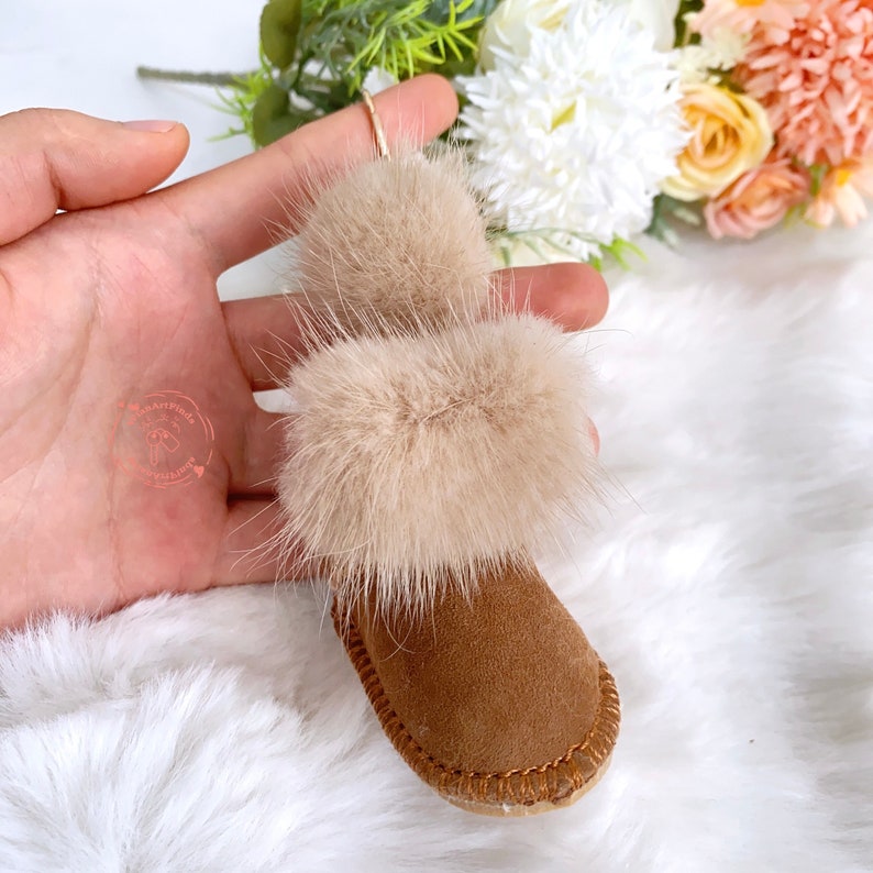 Puede incluir: Una peque&ntilde;a bota de gamuza marr&oacute;n con un ribete de piel blanca esponjosa y un pomp&oacute;n. La bota se sostiene en una mano.